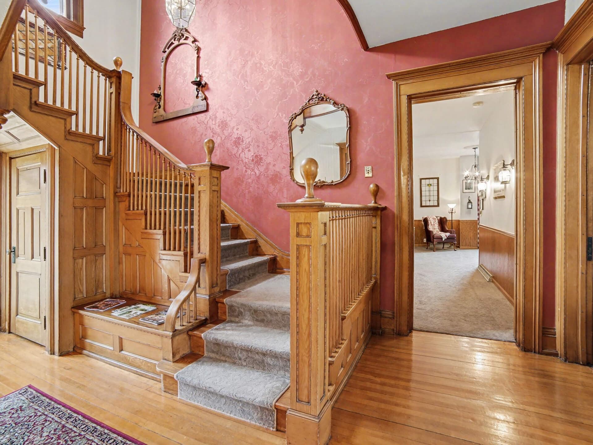 Elegant entryway with a wooden staircase, pink walls, and a doorway leading to a cozy living area.