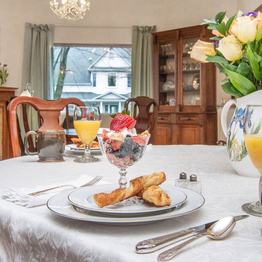 A beautifully set dining table featuring a fruit parfait, pastries, and drinks, with a floral centerpiece and a window view in the background.